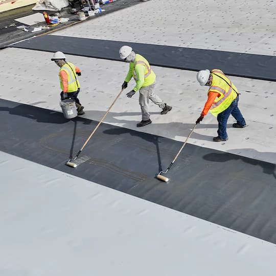 Three construction workers in safety gear apply adhesive to a flat roof using long rollers.